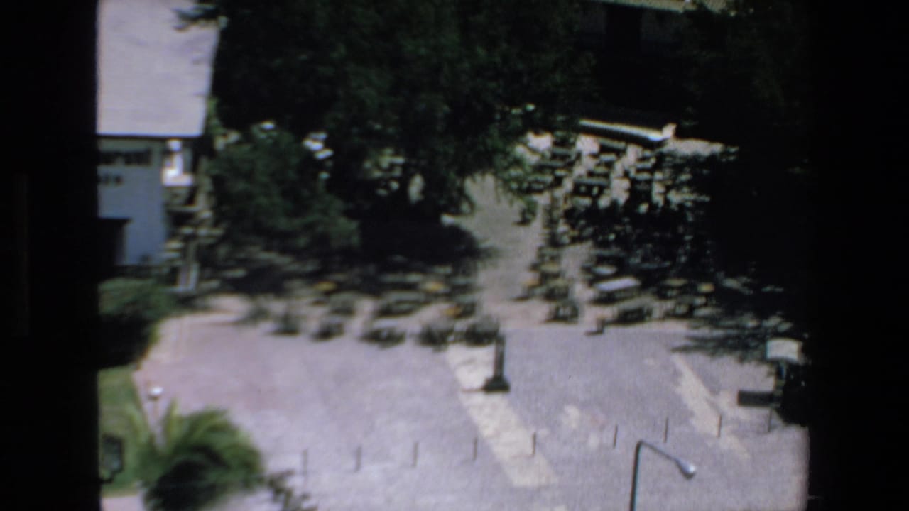 High-angle 8mm frame from Mazatlan showing parked cars and possible buses near shade trees, a white building, and open pavement.