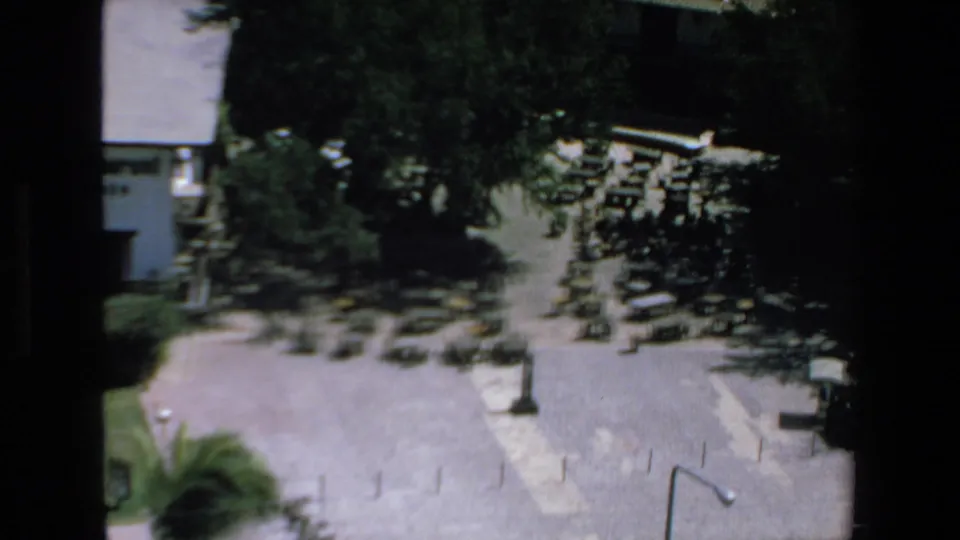 High-angle 8mm frame from Mazatlan showing parked cars and possible buses near shade trees, a white building, and open pavement.