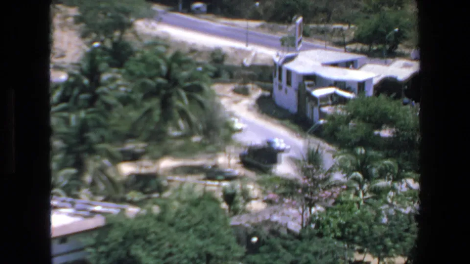 Elevated Mazatlan home-movie frame with a dark military-style truck, nearby cars, palm trees, roadways, and white buildings in a tropical setting.
