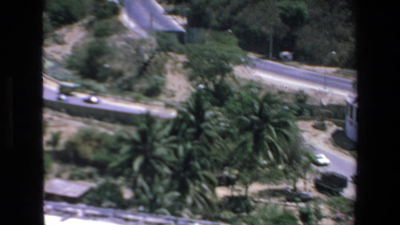 1972 Mazatlan footage shows an elevated view of a winding paved road bordered by palms and greenery, with small cars moving through the scene and dark framing at the edges.