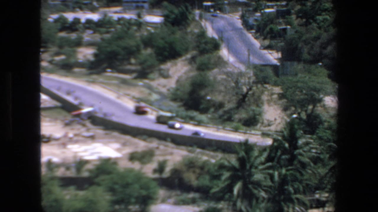 An elevated 1972 Mazatlan view looks over a winding road, scattered cars, palms, low vegetation, and sandy open ground framed by dark vertical borders.