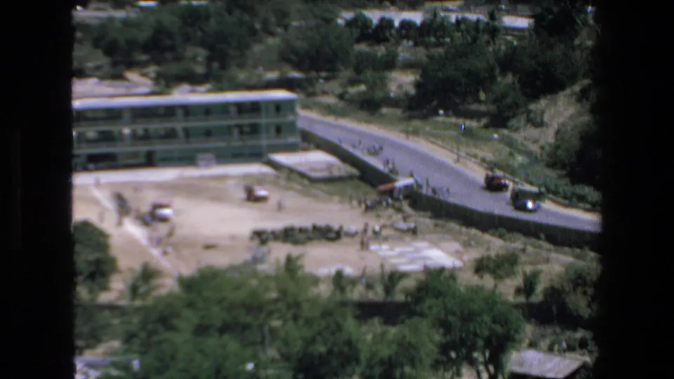 1972 Mazatlan footage shows a long multi-story building near a sandy field and road, with cars, trees, palms, and tiny distant people visible near the open ground.
