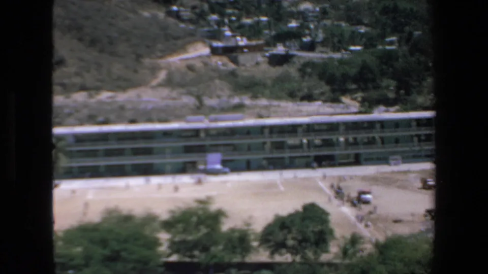 The 1972 Mazatlan view includes a long building beside sandy open ground, hillside roads, scattered vehicles, trees, and dark borders from the filmed vantage point.