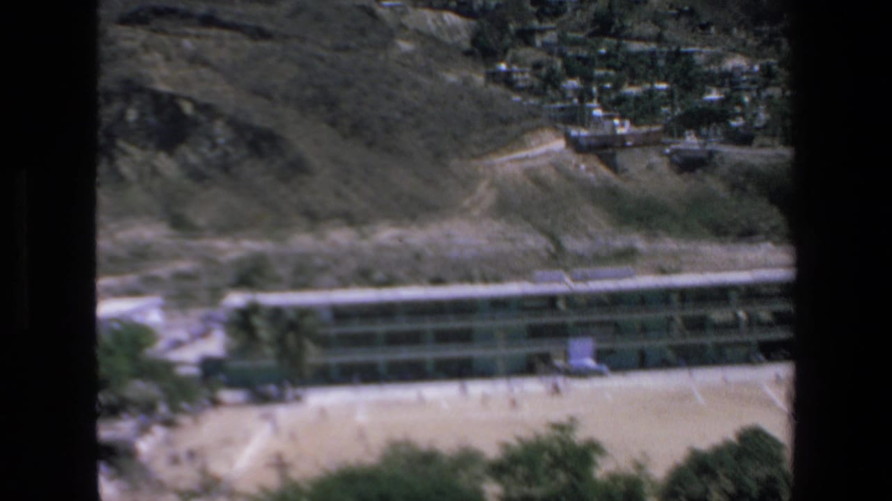 1972 Mazatlan home-movie frame showing a long rectangular hotel building at the base of a rugged hillside, with sandy open ground and small people visible near the structure.