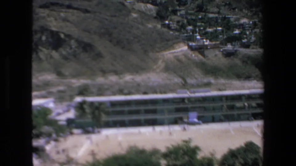 1972 Mazatlan home-movie frame showing a long rectangular hotel building at the base of a rugged hillside, with sandy open ground and small people visible near the structure.