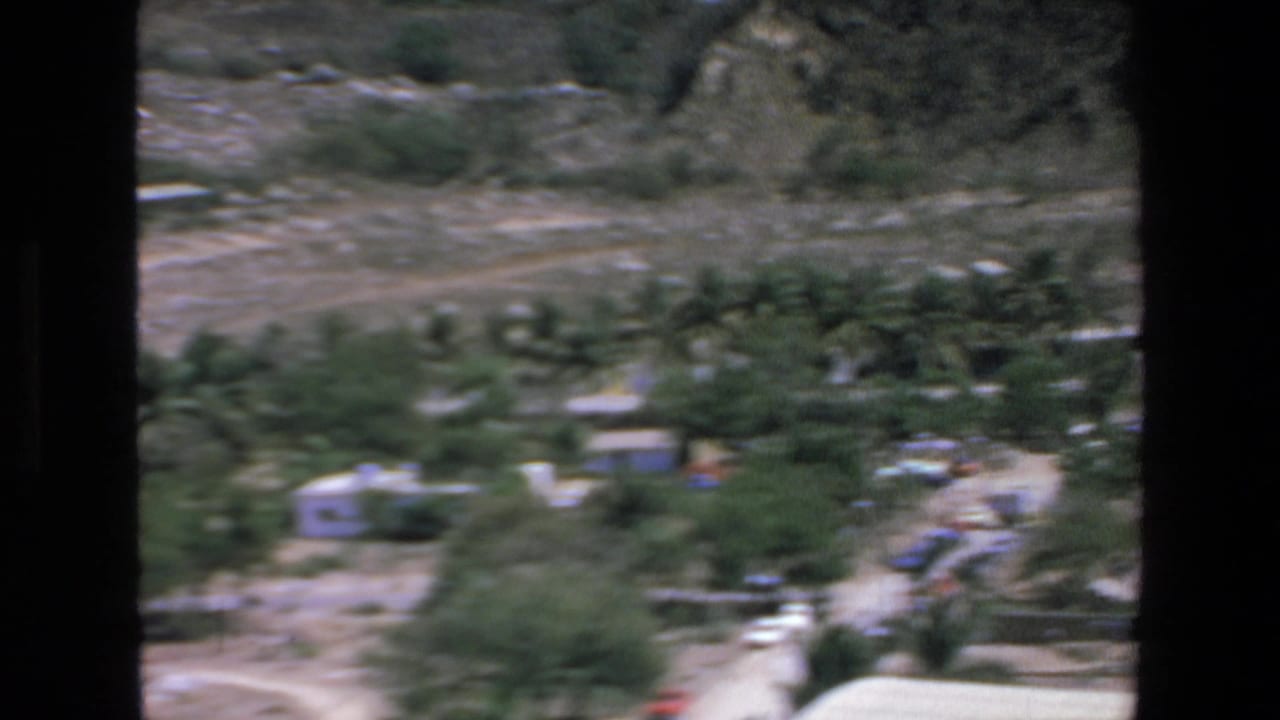 Blurred hillside resort area with palms, low buildings, dirt paths, and dry terrain.