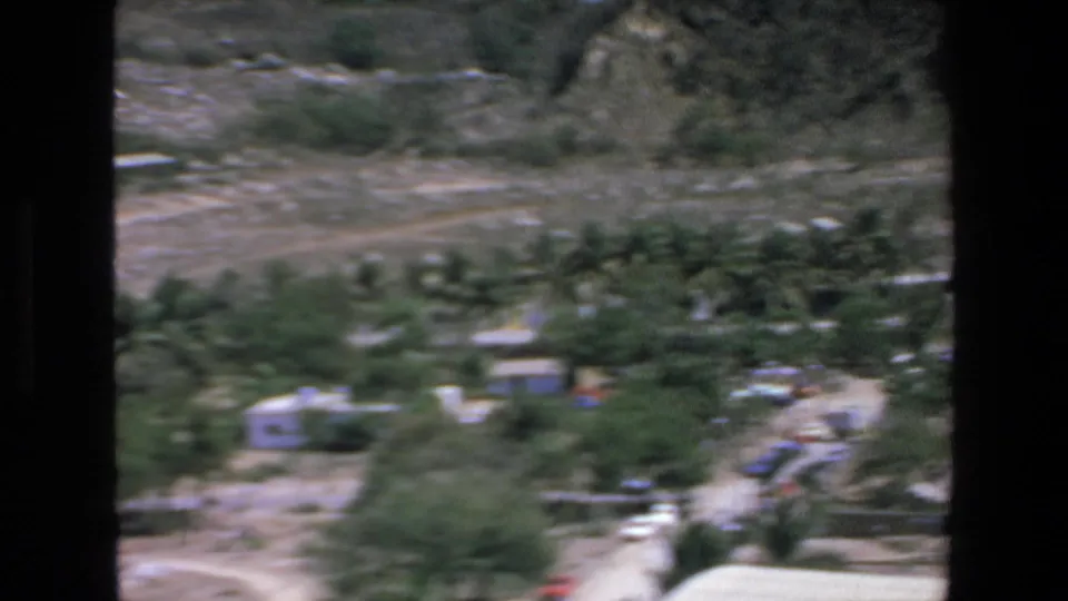 Soft 8mm frame from Mazatlan showing palms, low buildings, dry open ground, and hillside terrain during a handheld pan across the resort area.