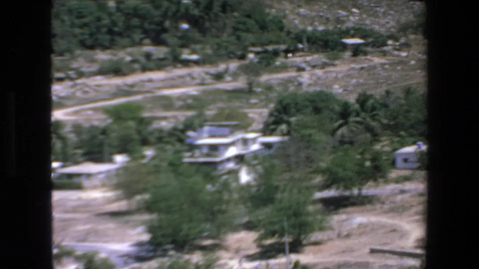 Elevated 1972 Mazatlan travel-film view across sandy resort grounds with trees, low structures, rough paths, and rocky hillside beyond.