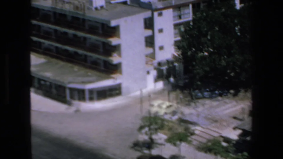 1972 Mazatlan home-movie frame showing the corner of a multi-story hotel with stacked balconies, a ground-floor entrance area, nearby trees, and open forecourt.