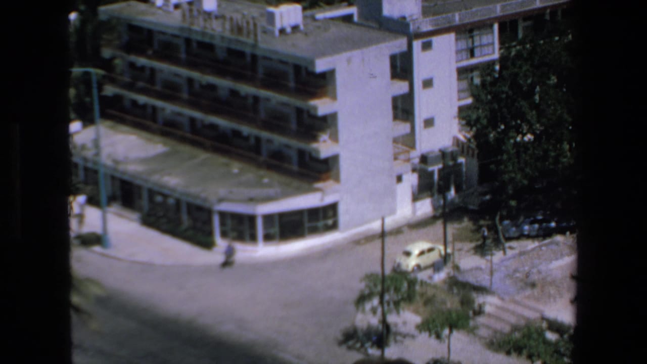 1972 Mazatlan home-movie frame showing an elevated view of a quiet street corner, modern low-rise buildings, trees, and a small yellow car.