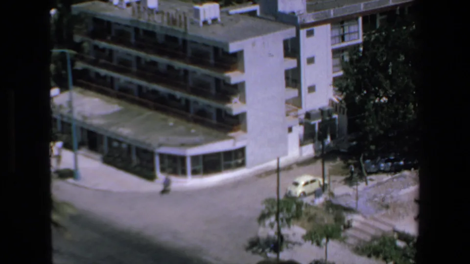 1972 Mazatlan home-movie frame showing an elevated view of a quiet street corner, modern low-rise buildings, trees, and a small yellow car.