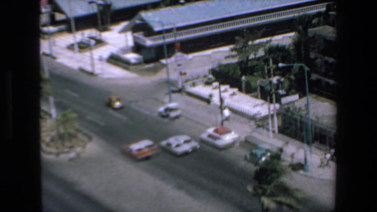 Elevated 1972 Mazatlan street view with several cars moving through an intersection, low buildings, sidewalks, and a small planted median.