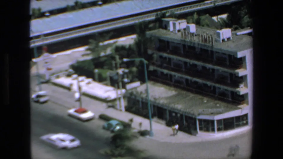 High-angle Mazatlan street frame showing older cars, a corner building with balconies, streetlights, and a bright sidewalk area.