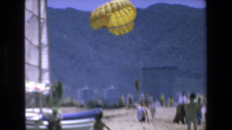 1972 Mazatlan waterfront frame with a yellow parachute drifting above the beach area, sailboat masts, distant mountains, water, and small human figures below.