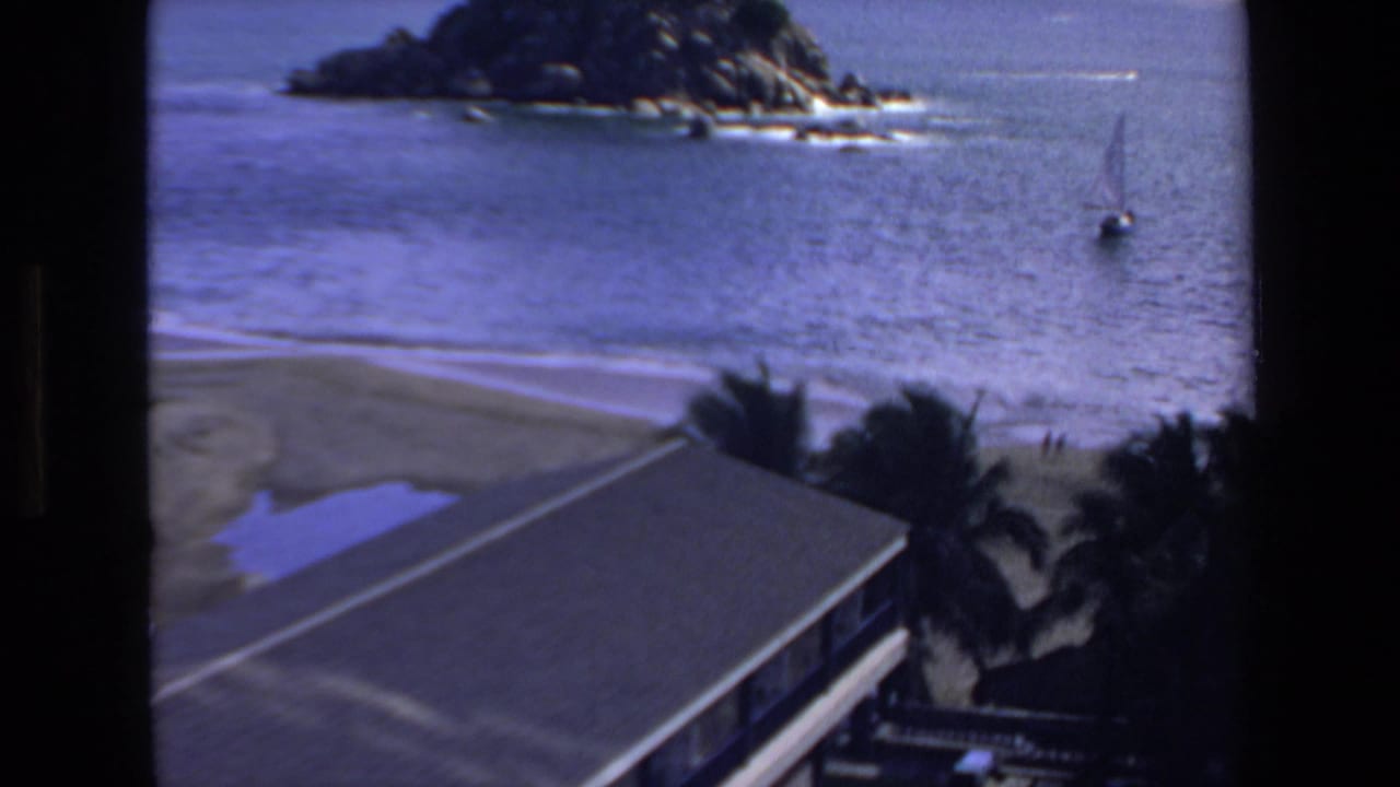 1972 Mazatlan home-movie frame showing a coastal beach view with a rocky offshore point, calm water, a small sailboat, palm trees, and a roof edge in the foreground.