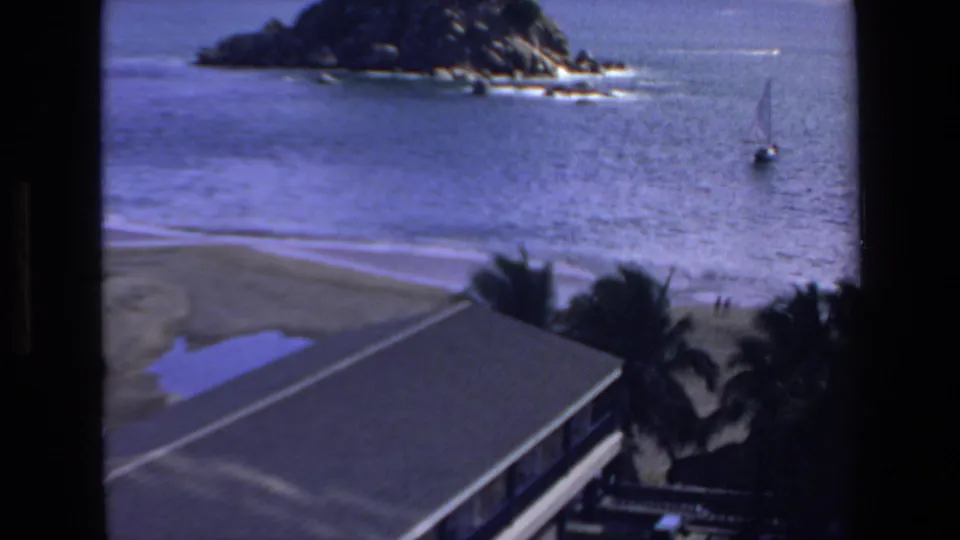 1972 Mazatlan home-movie frame showing a coastal beach view with a rocky offshore point, calm water, a small sailboat, palm trees, and a roof edge in the foreground.