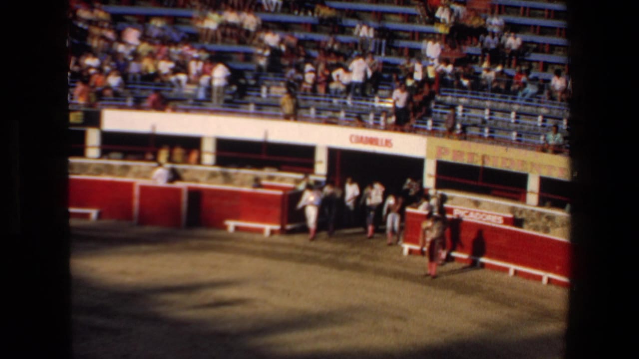 1972 Mazatlan bullring frame showing spectators in tiered seating as bullfighters or arena participants gather near the entrance below Spanish signage.