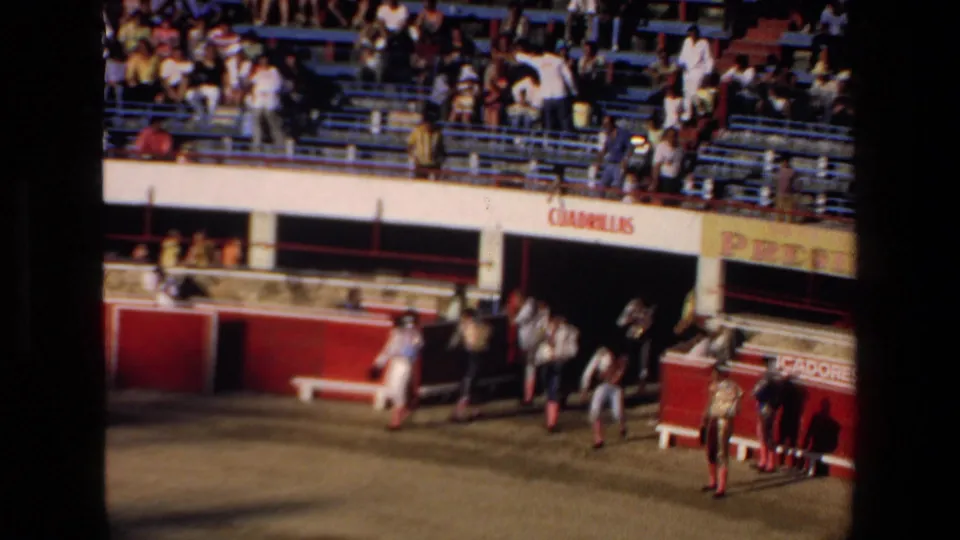 1972 Mazatlan home-movie frame showing a wider bullring view with spectators in the stands, red barriers, and bullfighters positioned near the entrance area.