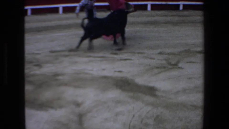 The black bull passes close to the matador and red cape in the sandy Mazatlan bullring, captured with strong motion blur and home-movie softness.