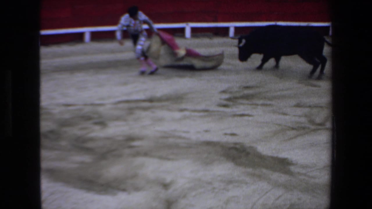 In supplied 1972 Mazatlan 8mm footage, a matador with a pink and gold cape faces a black bull inside a red-walled bullring.