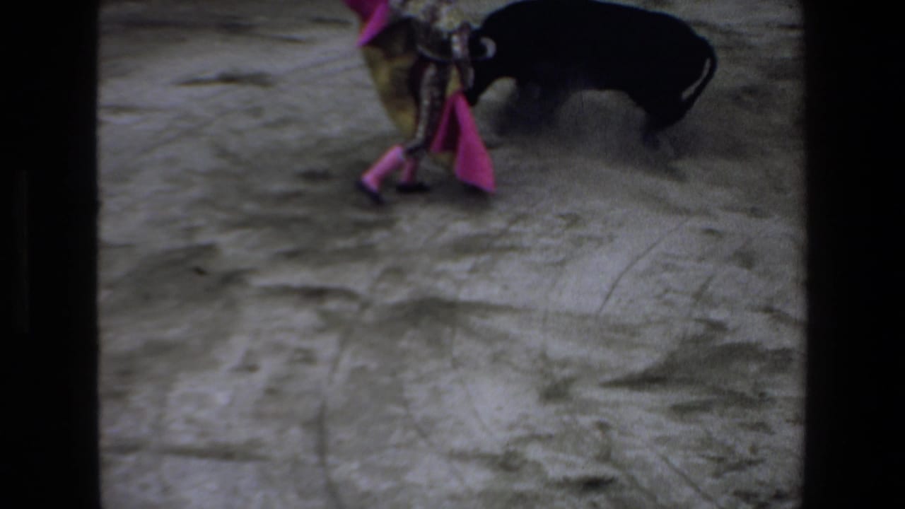 1972 Mazatlan home-movie frame showing a matador in a colorful costume moving a pink cape near a black bull in a dusty bullring.
