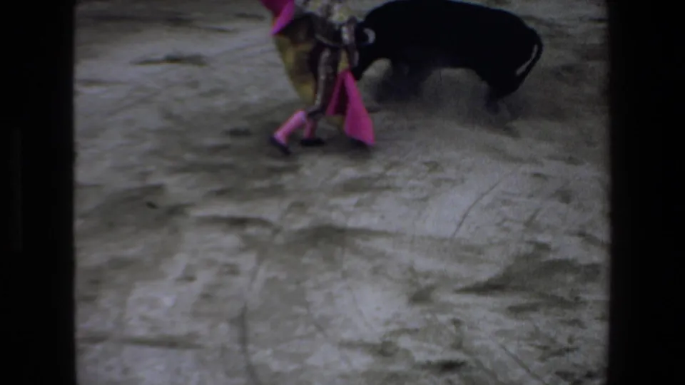 1972 Mazatlan home-movie frame showing a matador in a colorful costume moving a pink cape near a black bull in a dusty bullring.