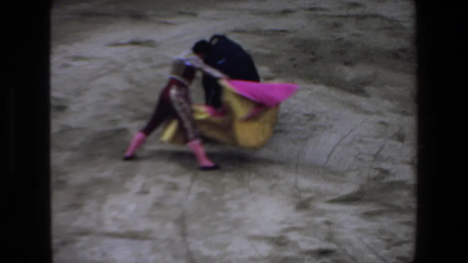 1972 Mazatlan frame showing the matador low to the ground with a pink and yellow cape while the black bull presses close in the arena.