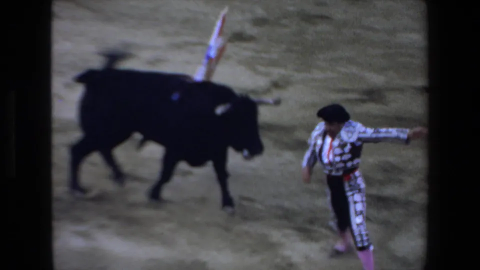 1972 Mazatlan home-movie frame showing a black bull with banderillas on its back near a matador in a decorated suit on dusty ground.