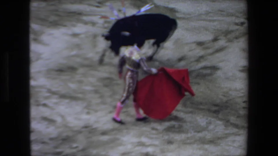 The matador holds a red cape in the sandy Mazatlan ring as the black bull turns nearby, with the handheld 8mm frame showing soft blur and dark borders.