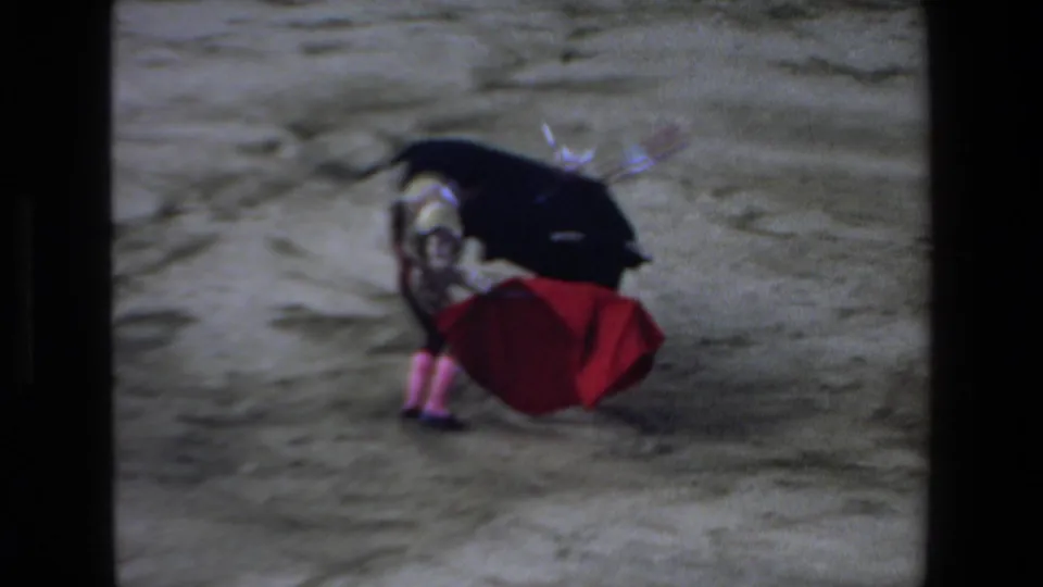 The matador gathers the red cape near a black bull marked with banderillas in the dusty ring, with home-movie film borders visible.