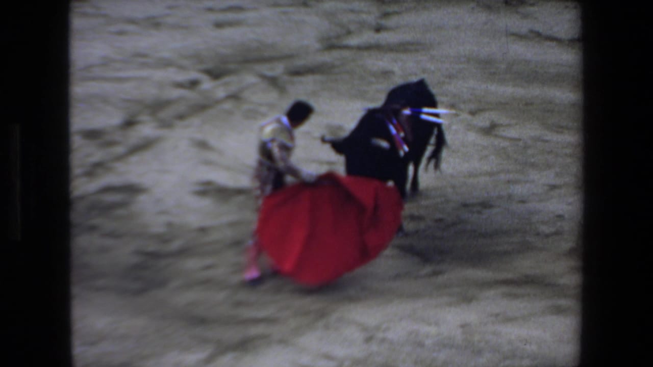 In metadata-supported 1972 Mazatlan footage, a matador in ornate dress maneuvers a red cape as a black bull presses close on sandy arena ground.