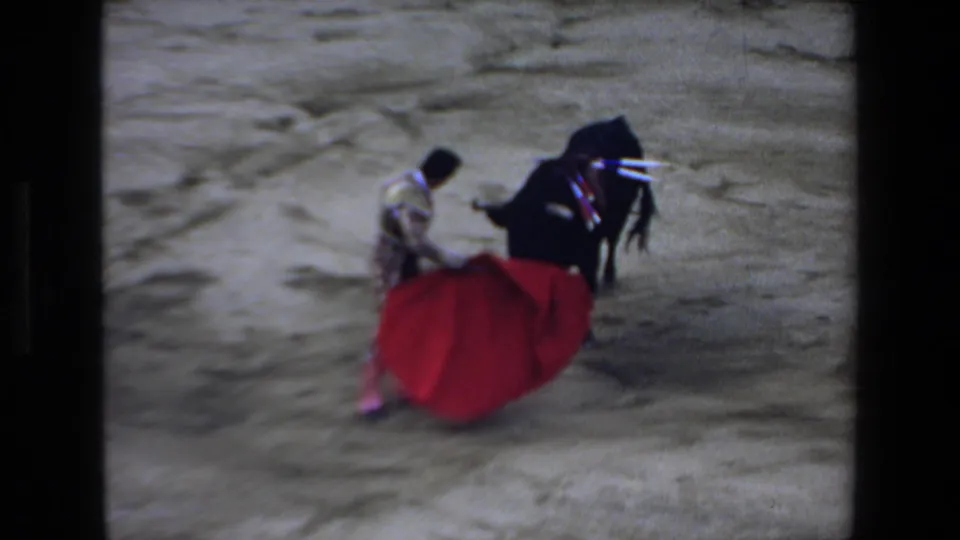 In metadata-supported 1972 Mazatlan footage, a matador in ornate dress maneuvers a red cape as a black bull presses close on sandy arena ground.