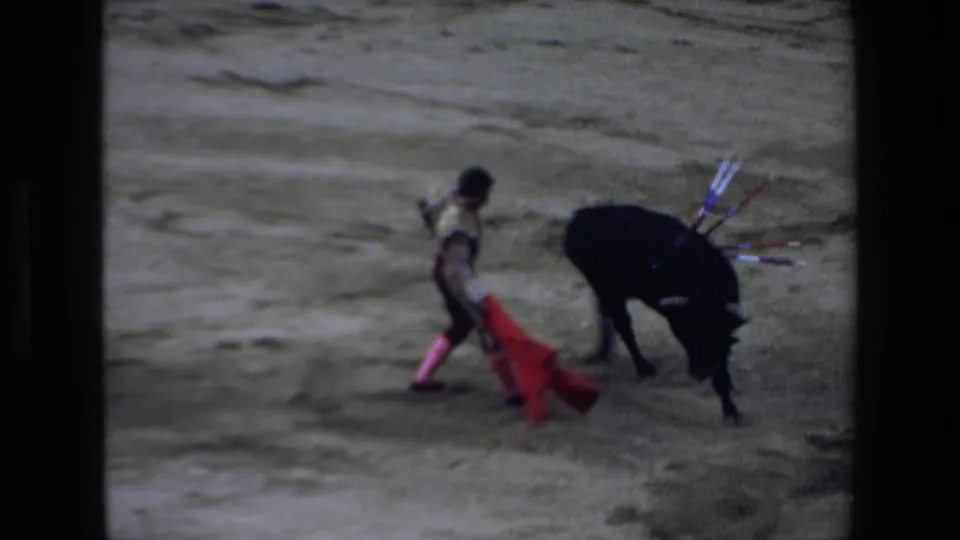 The matador moves across the arena with the red cape while the black bull turns behind him, captured in soft, grainy 8mm color.