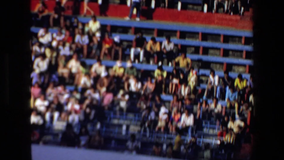 A crowd of spectators fills blue and red stadium benches, providing a brief view of the public audience around the bullfight arena.