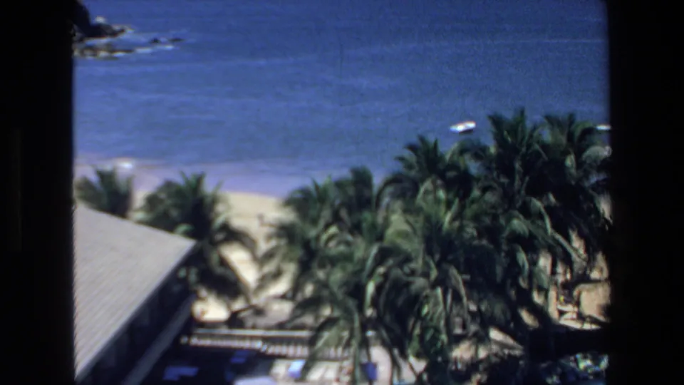 1972 Mazatlan resort view from above with palm trees, blue ocean, a beach edge, shaded structures, and a swimming pool area in the foreground.