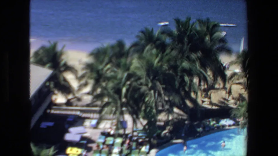 1972 Mazatlan home-movie frame looking over a resort swimming pool, palm trees, beach frontage, and blue ocean in bright daylight.