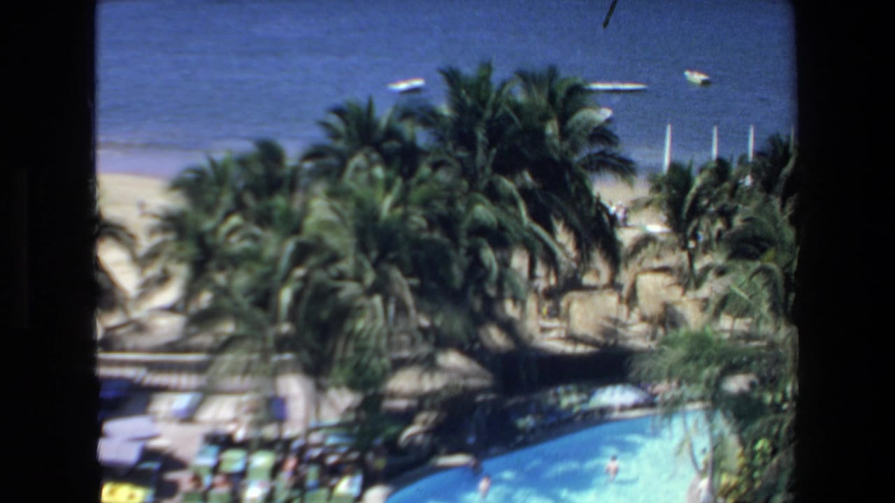 1972 Mazatlan, Mexico 8mm frame showing an elevated resort view with a curved turquoise pool, palm trees, beach, blue ocean, and small boats offshore.
