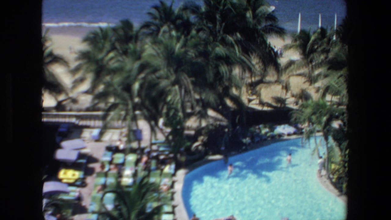 Resort pool and palm-lined beach area with ocean water and small boats in the background.