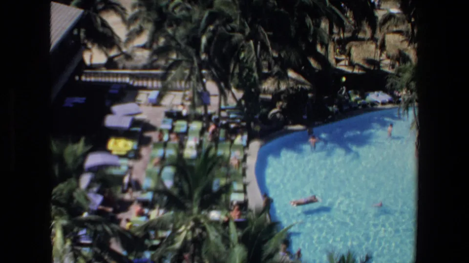 1972 Mazatlan home-movie frame showing swimmers in a curved resort pool, palm trees, umbrellas, lounge chairs, and the beach just beyond the deck.