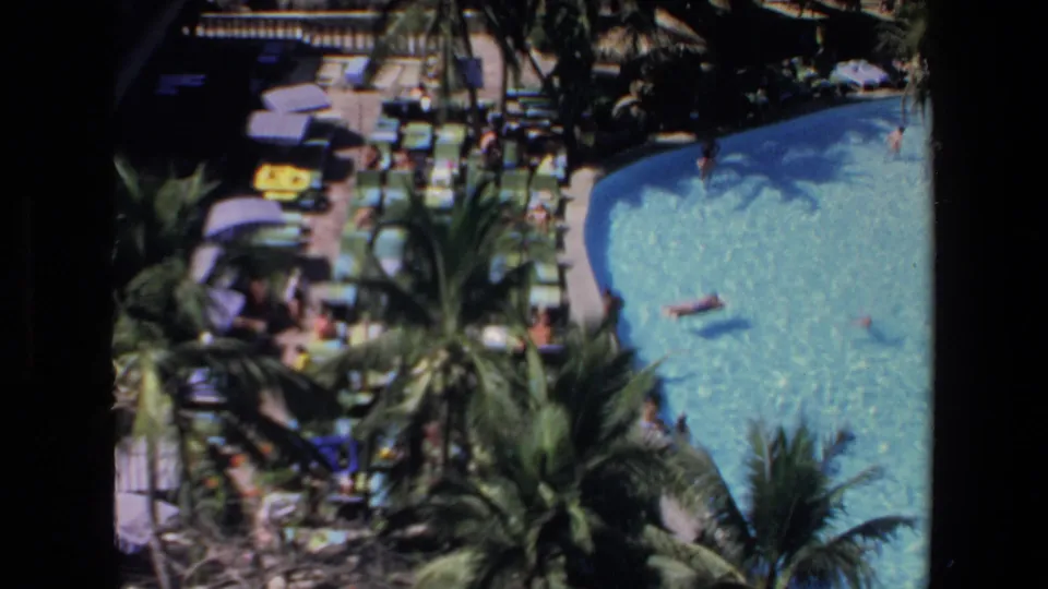 Elevated 1972 Mazatlan resort shot with a bright curved pool, distant swimmers, palm trees, umbrellas, poolside seating, and beach area near the ocean.