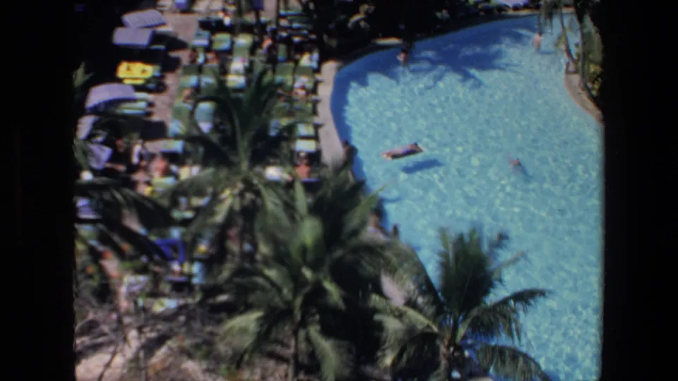 High-angle 1972 Mazatlan vacation view of a turquoise resort pool bordered by palms, lounge seating, umbrellas, swimmers, and inflatable rafts.