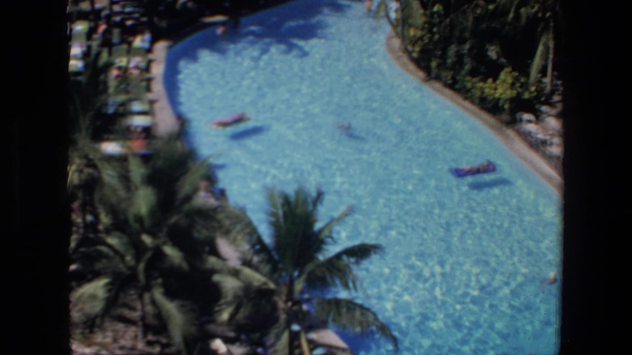 Elevated view of a bright blue pool with rafts, swimmers, palms, and rows of deck chairs.