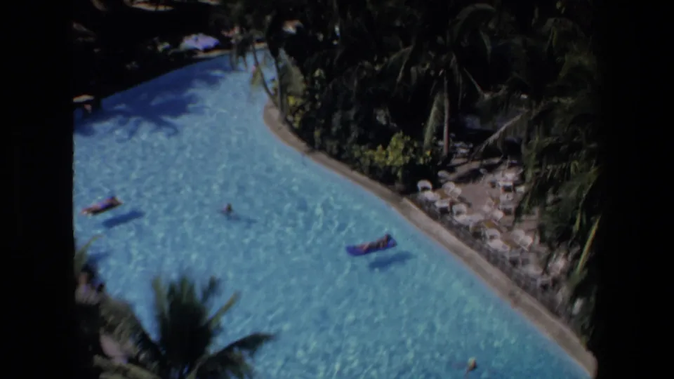 1972 Mazatlan home-movie frame of a curving resort pool section with drifting rafts, a swimmer, palm fronds, and landscaped planting beside the water.