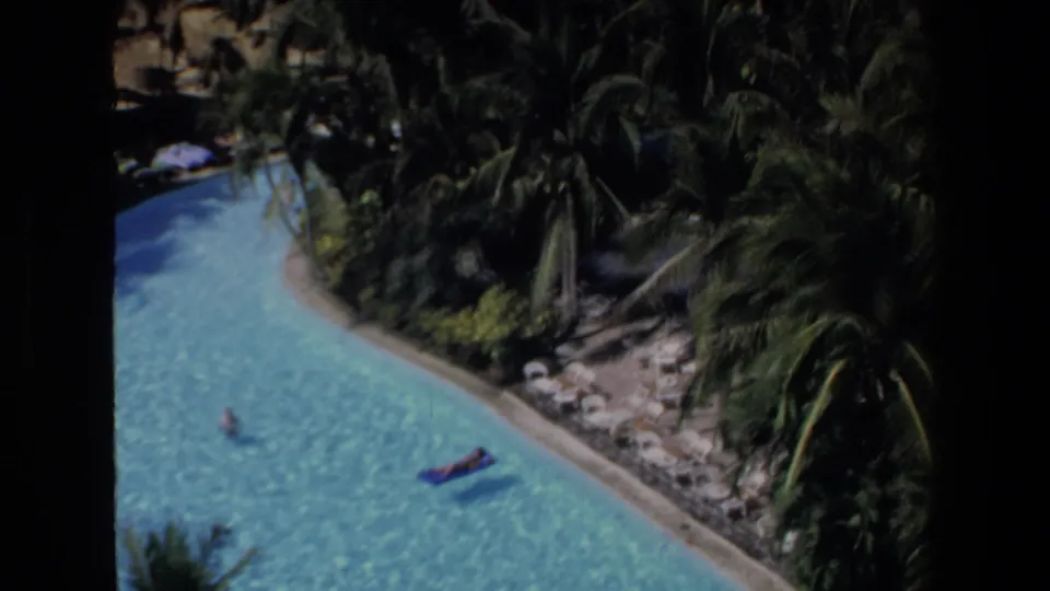 High-angle 1972 Mazatlan vacation frame showing a shaded corner of the resort pool with a raft, swimmers, palms, rocks, and tropical landscaping.