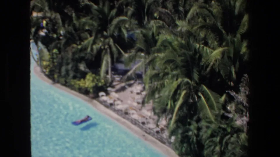 1972 Mazatlan home-movie frame showing a high-angle view of a curved turquoise resort pool bordered by palms, with a distant person on a blue float.