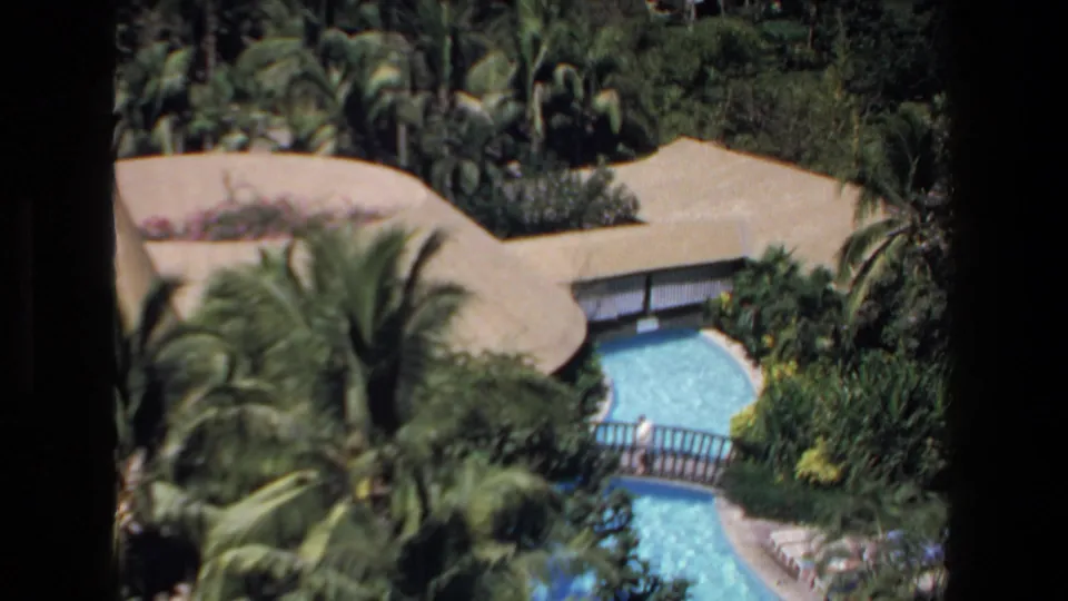1972 Mazatlan resort frame showing a winding turquoise pool crossed by a small bridge, with palms, walkways, and a light-colored roof or patio area nearby.