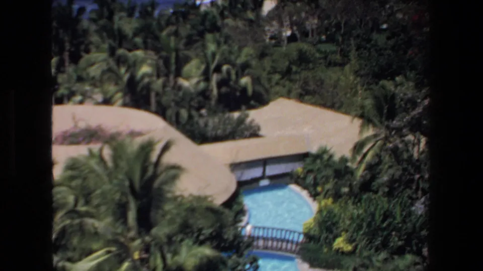 1972 Mazatlan home-movie frame looking down over palms, thatched resort structures, and a curved blue swimming pool.