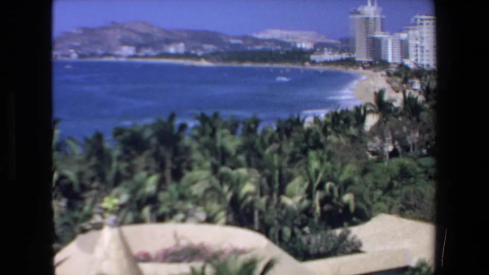1972 Mazatlan home-movie view across a blue bay toward a sandy beach, high-rise hotels, palms, and resort rooftops.