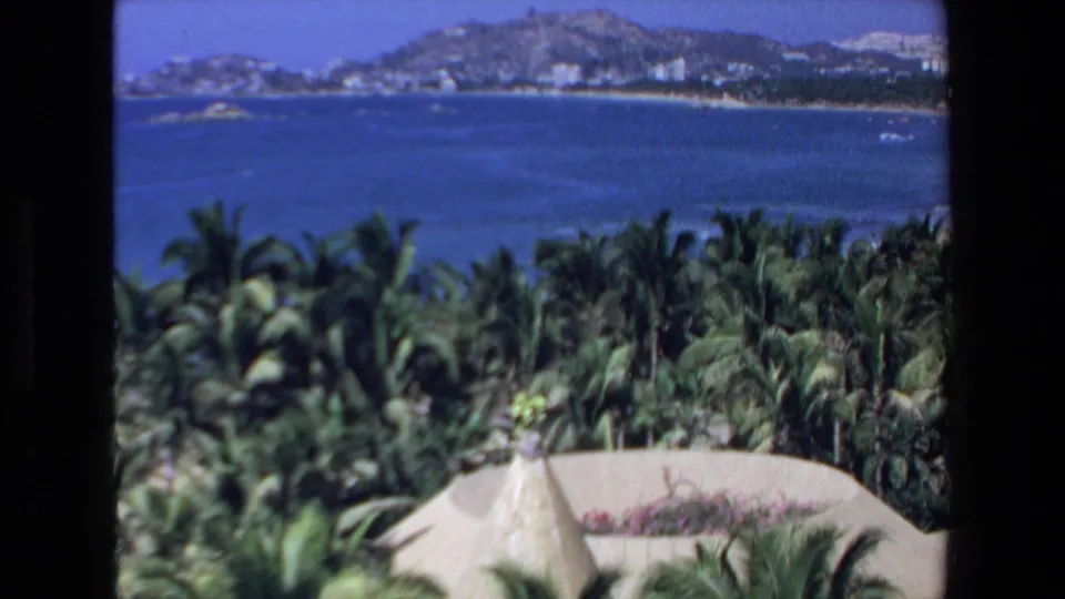 1972 Mazatlan resort overlook showing ocean water, palm canopy, a thatched roof, beach strip, and low hills in the distance.
