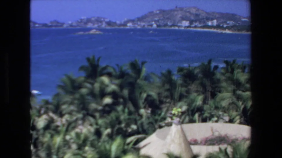 1972 8mm view from Mazatlan, Mexico, looking over palms toward a blue harbor, distant hills, and white waterfront buildings.